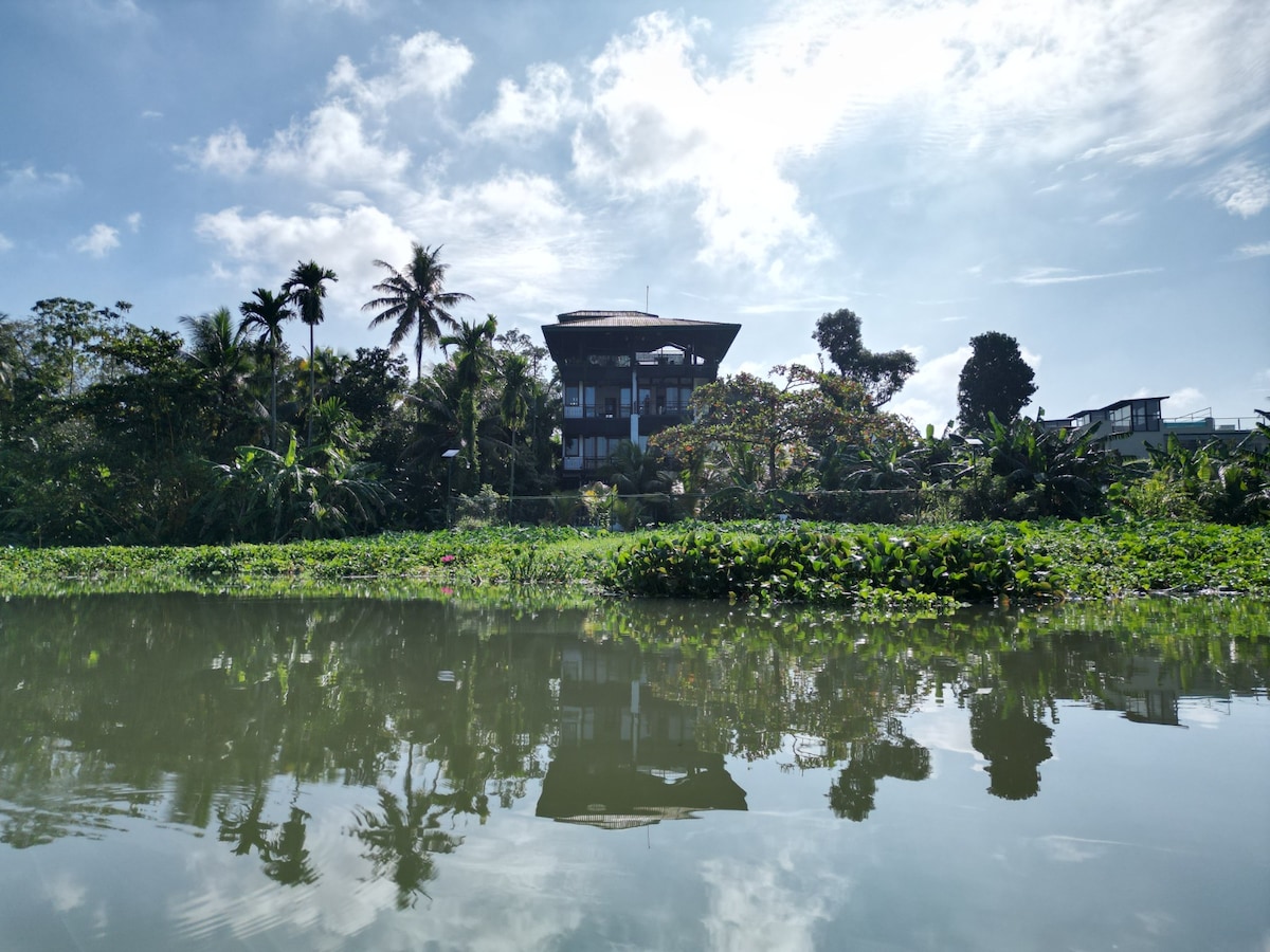 A modern multi-story building is surrounded by lush greenery, reflecting in the calm river water. Tall trees and vibrant plants are visible, contributing to a serene environment. The sky is bright with puffy clouds, enhancing the natural setting of the location.