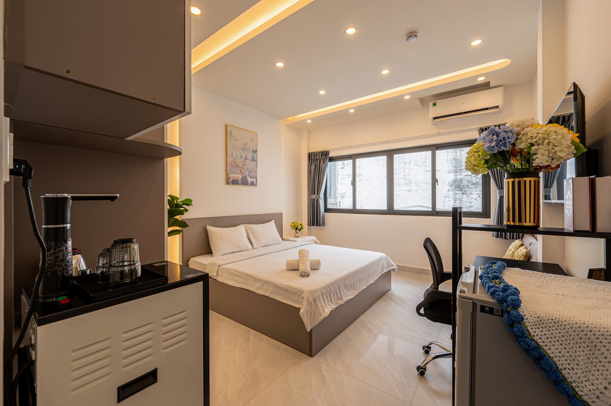 A bright and airy superior room features a spacious bed with soft bedding. Large windows allow natural light, and a kitchenette area is visible in the corner. Modern decor is complemented by a small plant and decorative flowers on a desk.