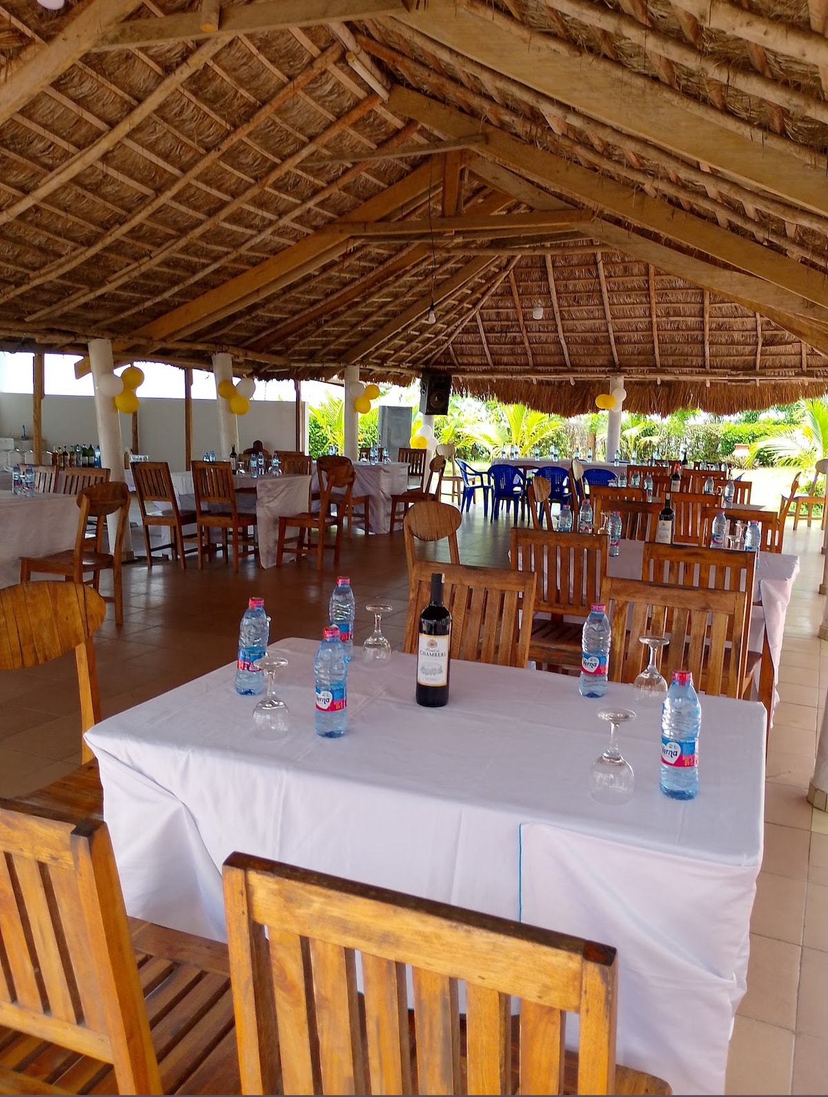 An open dining area is showcased, featuring wooden tables set with glasses and water bottles. The ceiling is adorned with natural materials, enhancing the atmosphere. Surrounding vegetation is visible, offering a sense of connection to the outdoors.
