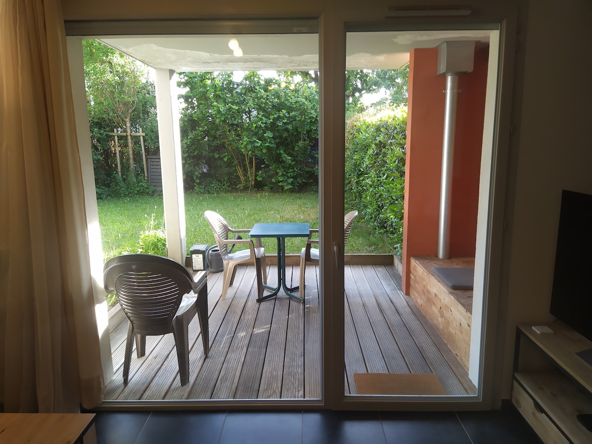 A view from inside the apartment shows a covered terrace with a small table and two chairs. Lush greenery surrounds the area, creating a private outdoor space. The wooden decking leads to a small step, providing access to the garden area beyond.