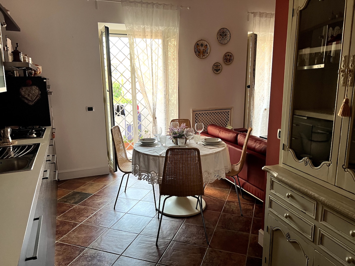 A dining area is featured, with a round table set for four, adorned with white dishes and a floral centerpiece. Two large windows allow natural light to fill the space, complemented by light curtains. A cozy sofa in rich red is positioned nearby.