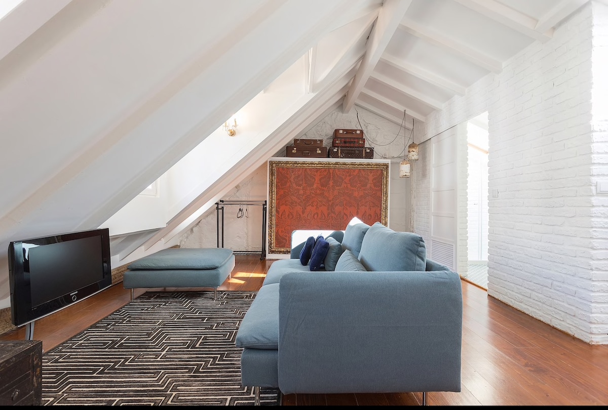 A cozy attic space features sloped ceilings with exposed beams. A blue sofa offers comfortable seating, complemented by a large floor cushion and a patterned rug. Antique luggage is displayed on the wall, while a minimalistic TV is positioned nearby, creating a relaxed atmosphere.