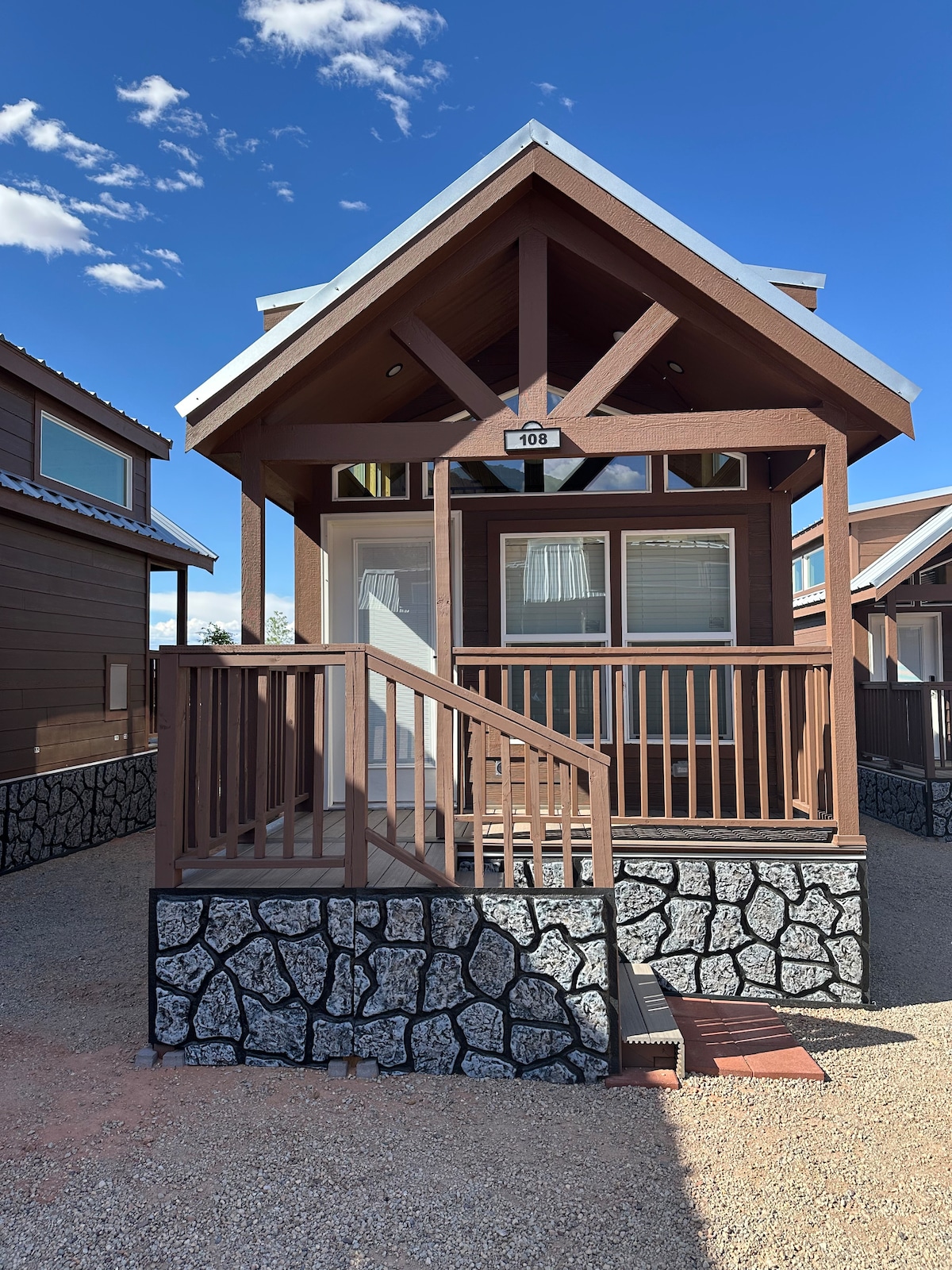 A charming cabin exterior is presented with a wooden façade and a peaked roof. A small covered porch with a railing is visible, along with steps leading to the entrance. Landscaping features gravel and stone elements, complementing the structure's natural surroundings.