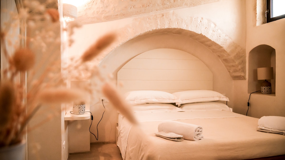 A spacious sleeping area features a large bed with soft linens, accompanied by two neatly rolled towels. The room is accentuated by stone archways and subtle recessed lighting, providing a calming ambiance. Minimalistic decor completes the serene atmosphere, with a bedside table and lamp visible.