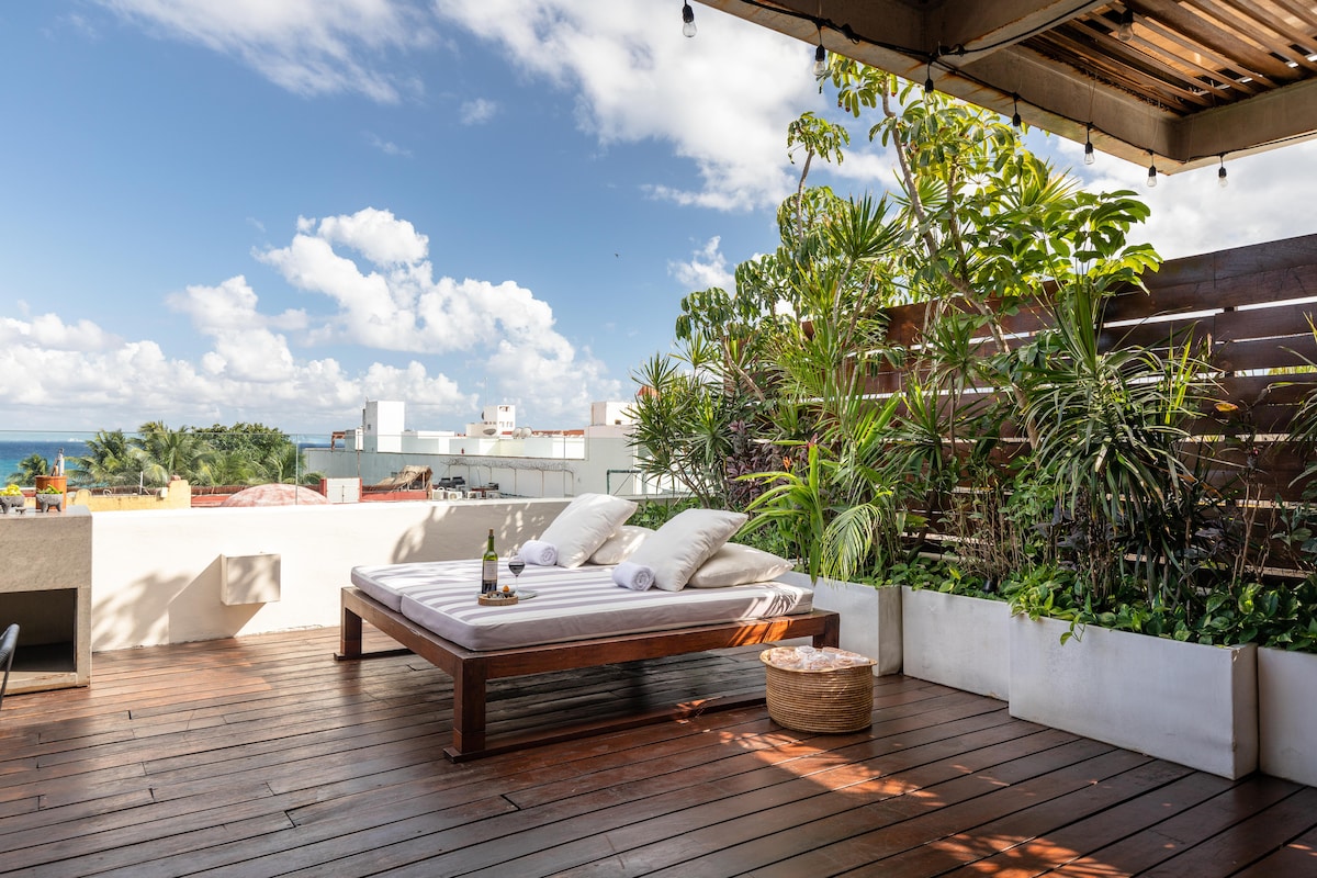 A rooftop terrace is presented, adorned with lush greenery and wooden decking. A spacious daybed, set with soft cushions, invites relaxation under a bright sky. A bottle of champagne sits elegantly beside the daybed, enhancing the tranquil atmosphere of the space.