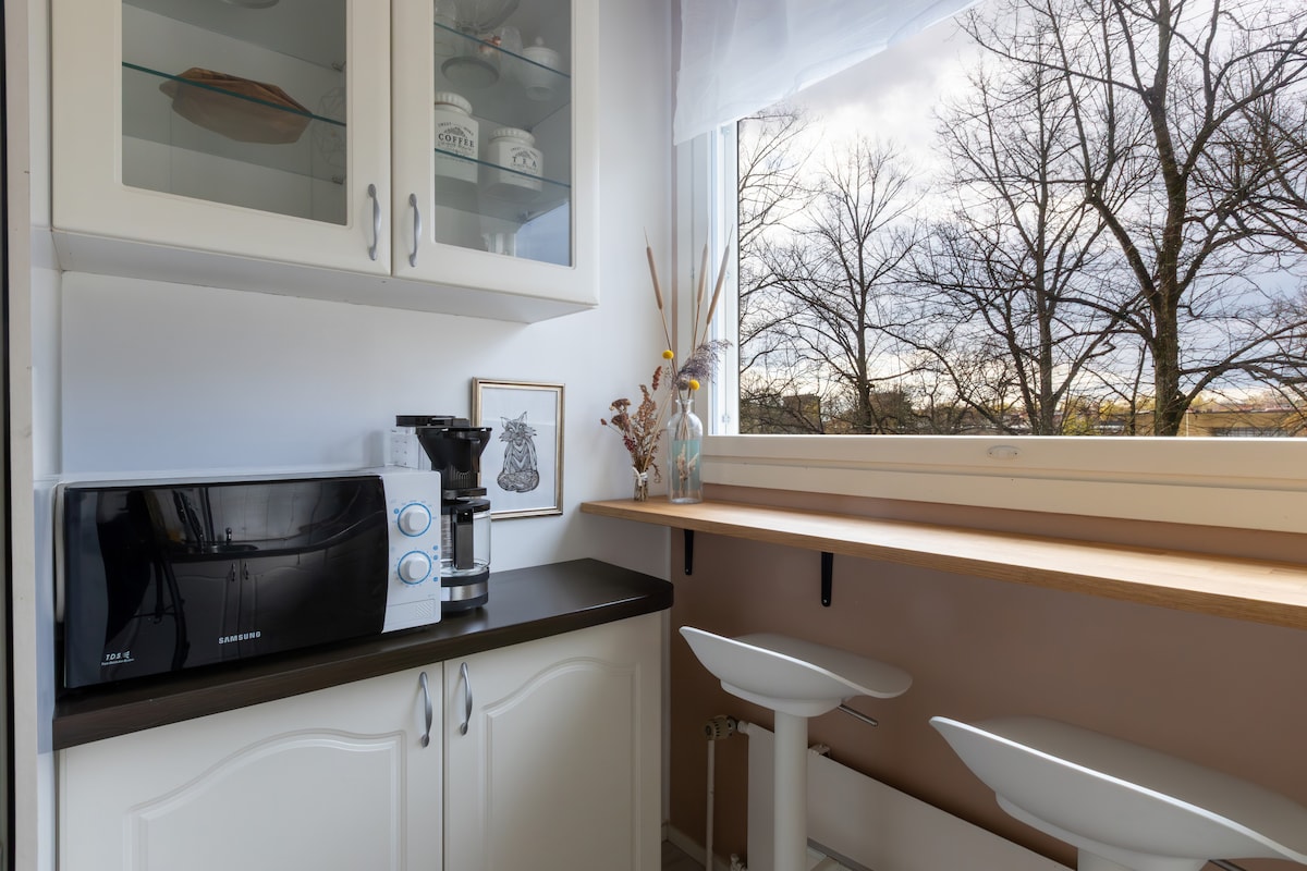 A kitchen corner features a microwave and a coffee maker on a dark countertop. Two white bar stools are positioned against a window, which offers a view of bare tree branches outside. A framed art piece and decorative elements add subtle interest to the space.