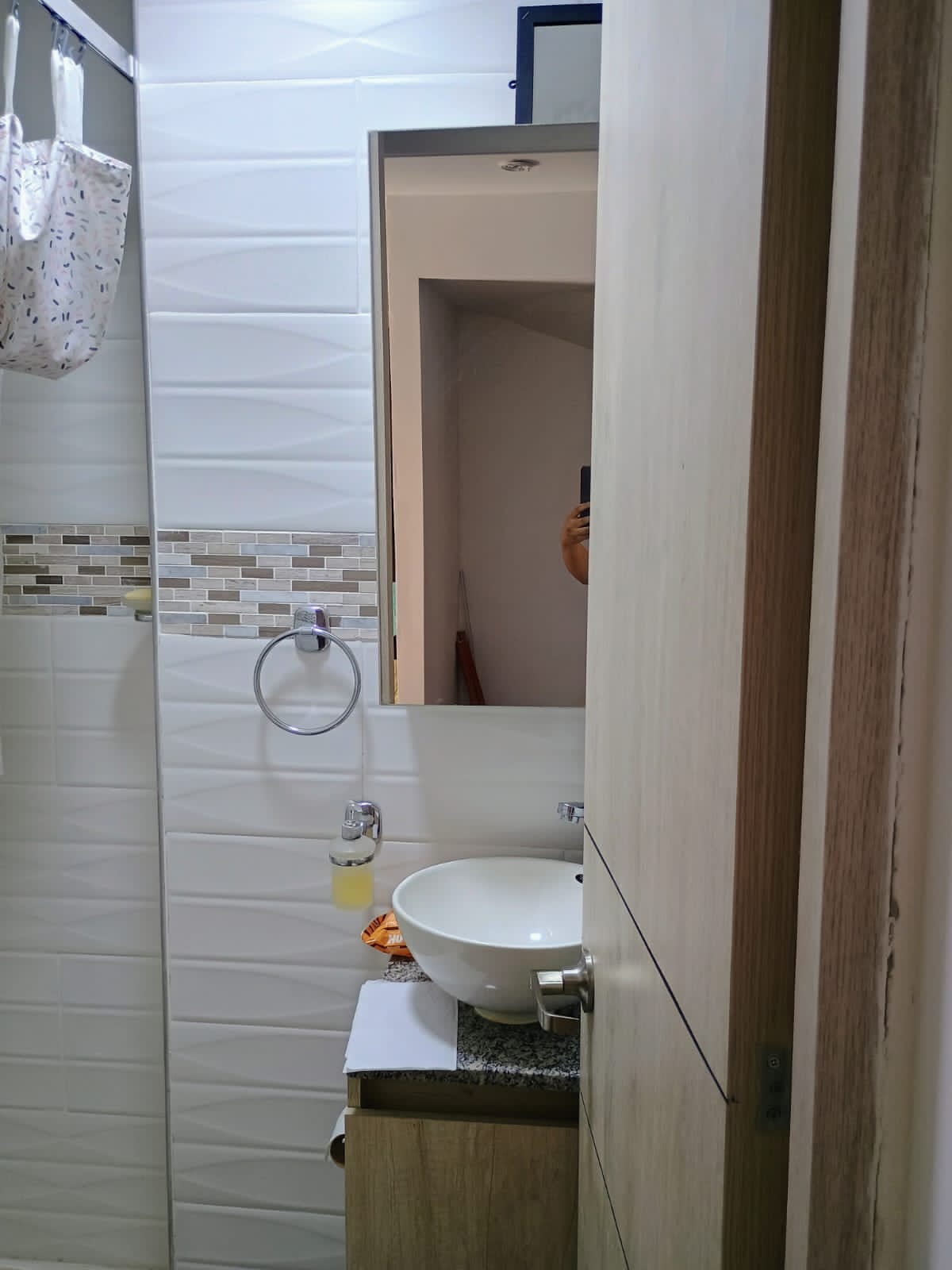 A modern bathroom is displayed, featuring a round basin atop a wooden vanity. A neatly mounted mirror reflects the space, with light tiles complemented by a decorative strip of earthy tones. A towel hangs on a hook, enhancing the bathroom's functional design.