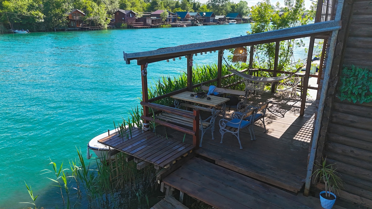 A wooden deck extends over clear blue water, featuring a dining table surrounded by chairs and a hammock hanging nearby. Lush green plants border the deck, and rustic wooden structures can be seen across the water, creating a serene outdoor space.