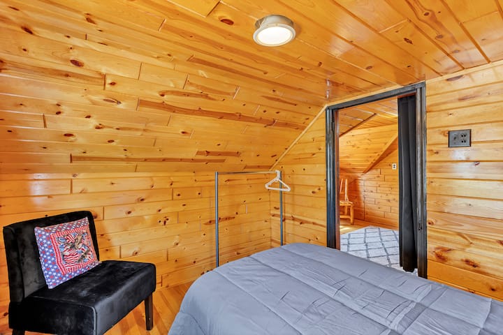 Este acogedor dormitorio tipo loft, situado bajo cálidos techos con paneles de madera, cuenta con una lujosa cama, iluminación suave y una elegante silla decorativa, perfecta para leer o relajarse en un refugio privado, similar a una cabaña.
