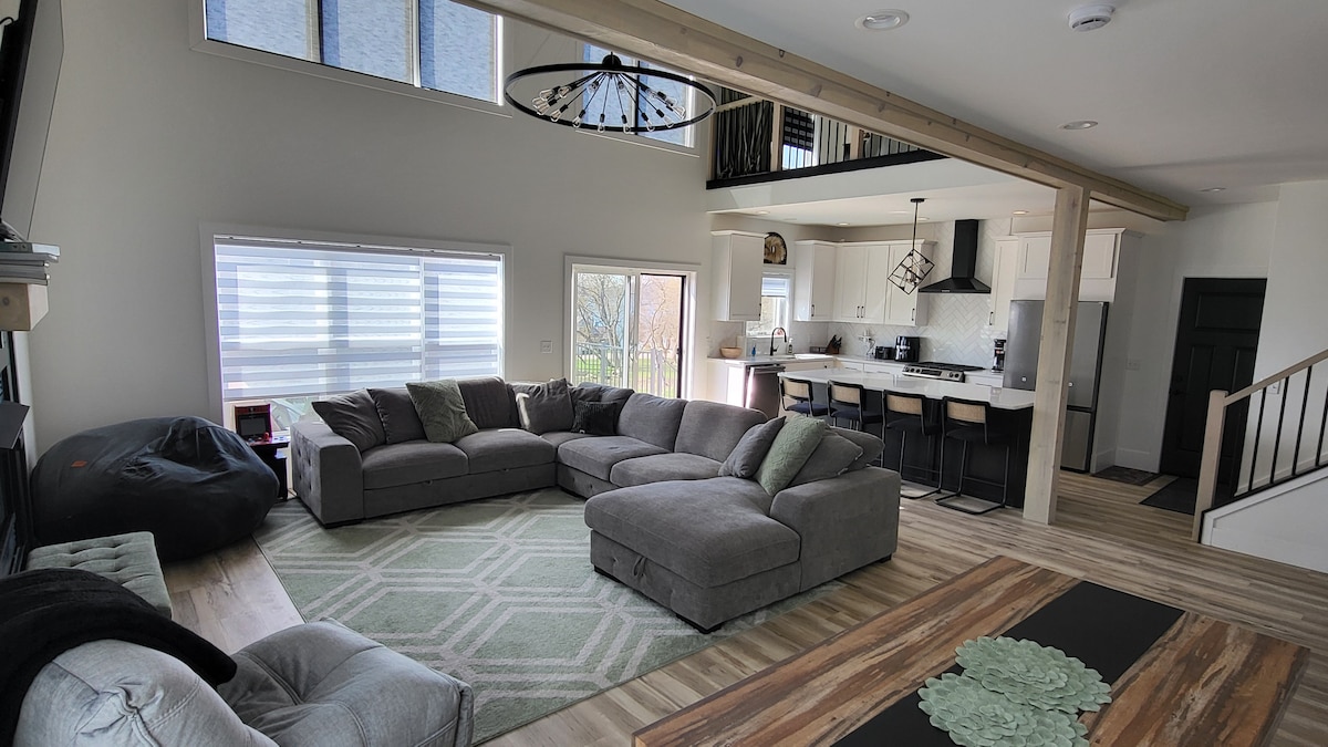 A spacious living area features a large sectional sofa positioned on a light-colored rug. Large windows allow natural light to fill the space, complementing the modern kitchen with an island in the background. An inviting dining area is visible, enhancing the communal atmosphere.