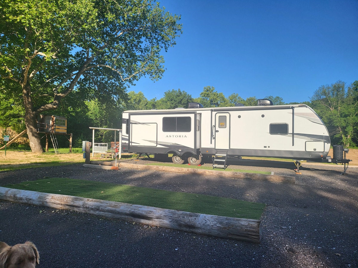 A modern camper trailer is parked on a gravel surface, surrounded by greenery. A nearby treehouse is visible on the left, and a small cart is positioned beside the camper. A grassy area is present in front, enhancing the outdoor setting.
