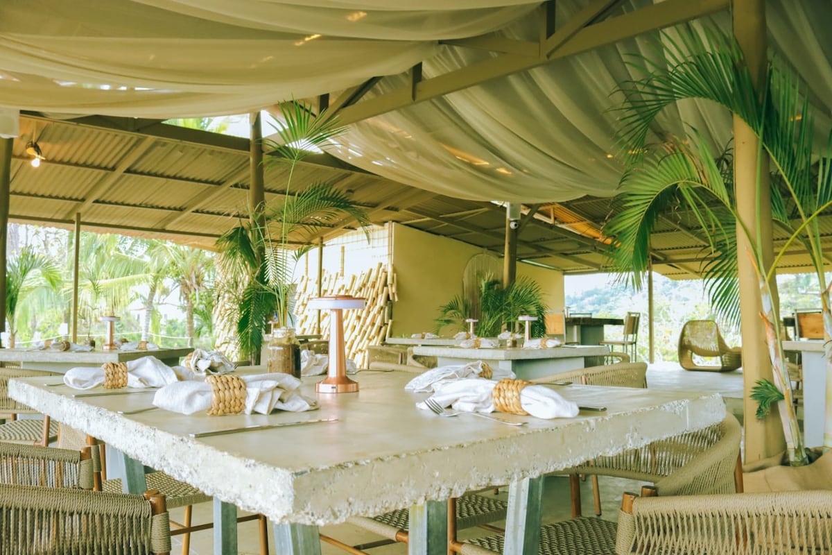 A dining area features several spacious tables, each set with neatly arranged napkins and utensils. Soft drapes overhead create a relaxed ambiance, complemented by tropical plants placed strategically throughout the space. Natural light fills the room, enhancing the open-air feel with views of the surrounding greenery.
