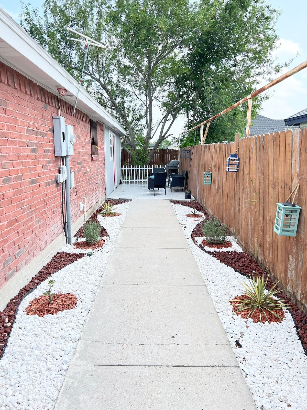 A concrete walkway is bordered by landscaped areas featuring a mix of white gravel and red mulch. Small plants and shrubs are positioned along the path. A pair of chairs are visible at the end of the walkway, surrounded by greenery and a wooden fence on one side.