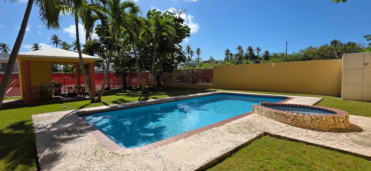 A swimming pool is surrounded by lush greenery and palm trees, with clear blue water reflecting the sunny sky. A seating area is visible nearby, providing a space for relaxation, while a stone-accented spa area is integrated into the pool design.
