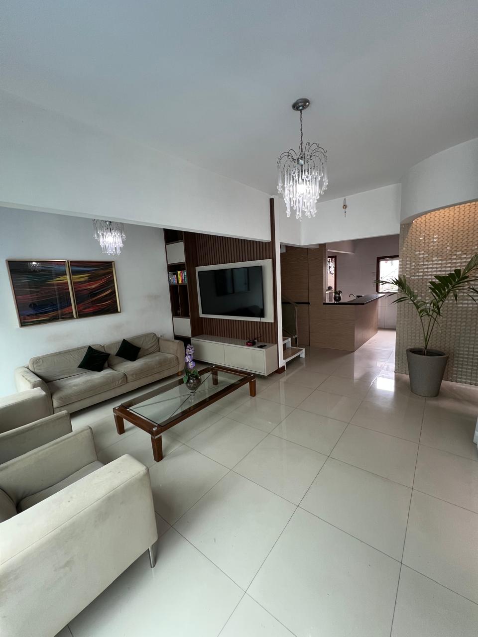 A modern living space features a cozy seating area with a glass coffee table and two light-colored sofas. Wall art adds a splash of color, while a large television is mounted for convenience. A potted plant brings a touch of greenery, and light fixtures provide soft illumination.