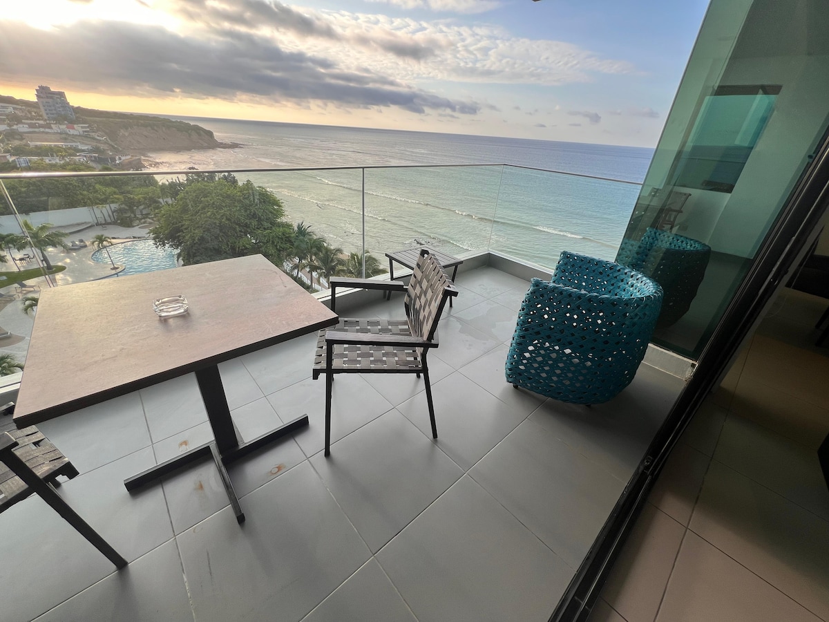 A spacious balcony overlooks the ocean, featuring a table and two chairs. One chair is designed in a bright blue color, while the other is a dark, sturdy material. The expansive view includes beach and waves, framed by delicate clouds above.