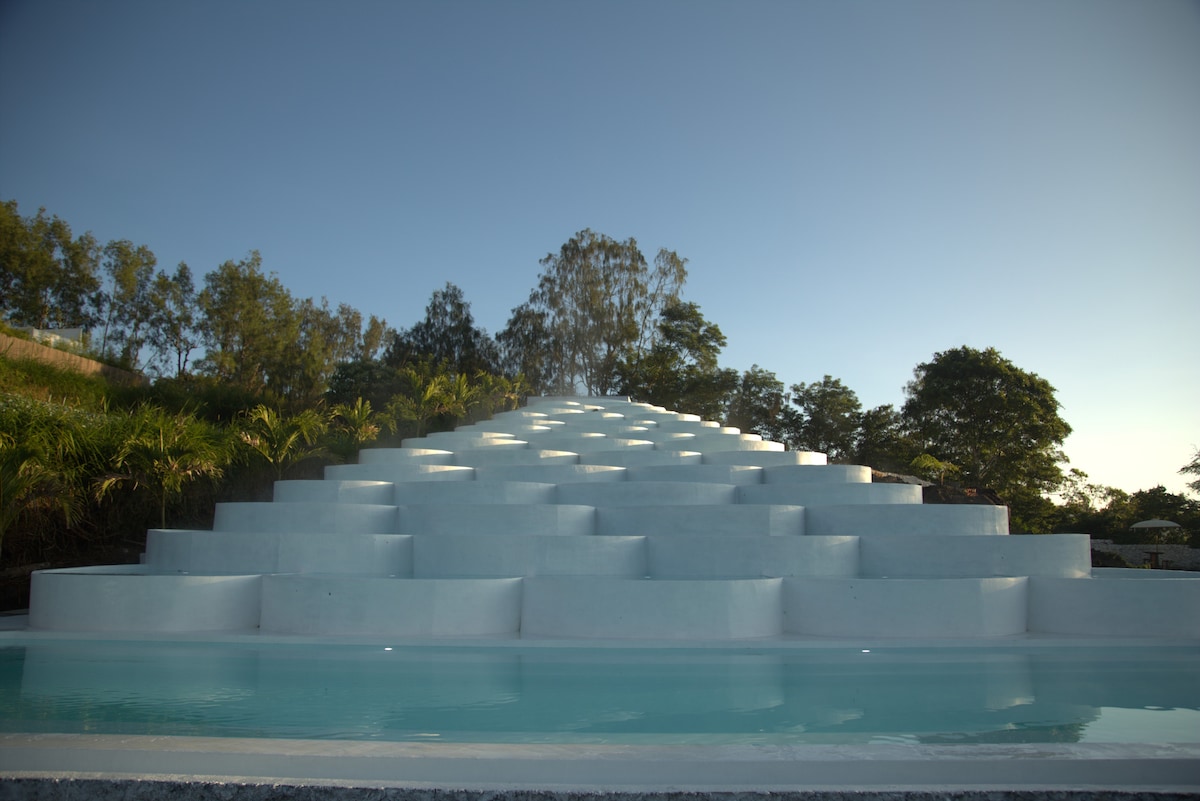 The unique Pyramid Infinity Hot Spring Pool is showcased, featuring a series of tiered steps leading to a tranquil water surface. Lush greenery surrounds the pool, illuminated by soft natural light and framed against a clear blue sky.