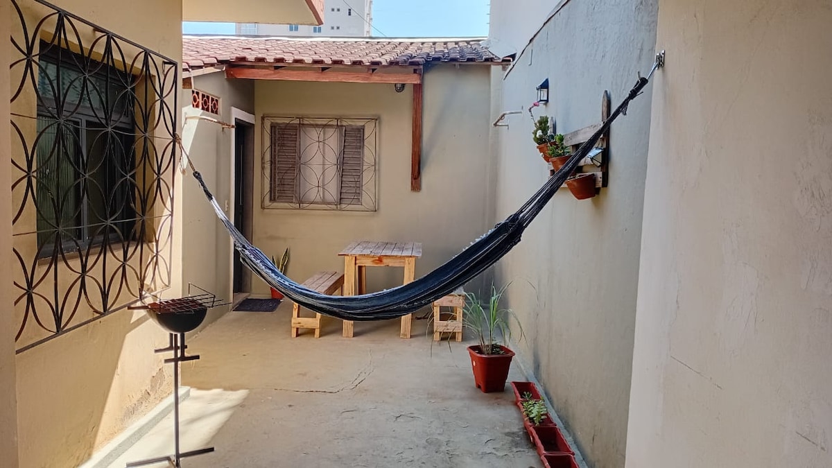 An outdoor patio space is shown, featuring a black hammock hung between walls. A wooden table sits beside it, accompanied by potted plants on the ground and mounted on the wall. A small grill is positioned nearby, enhancing the area for relaxation.