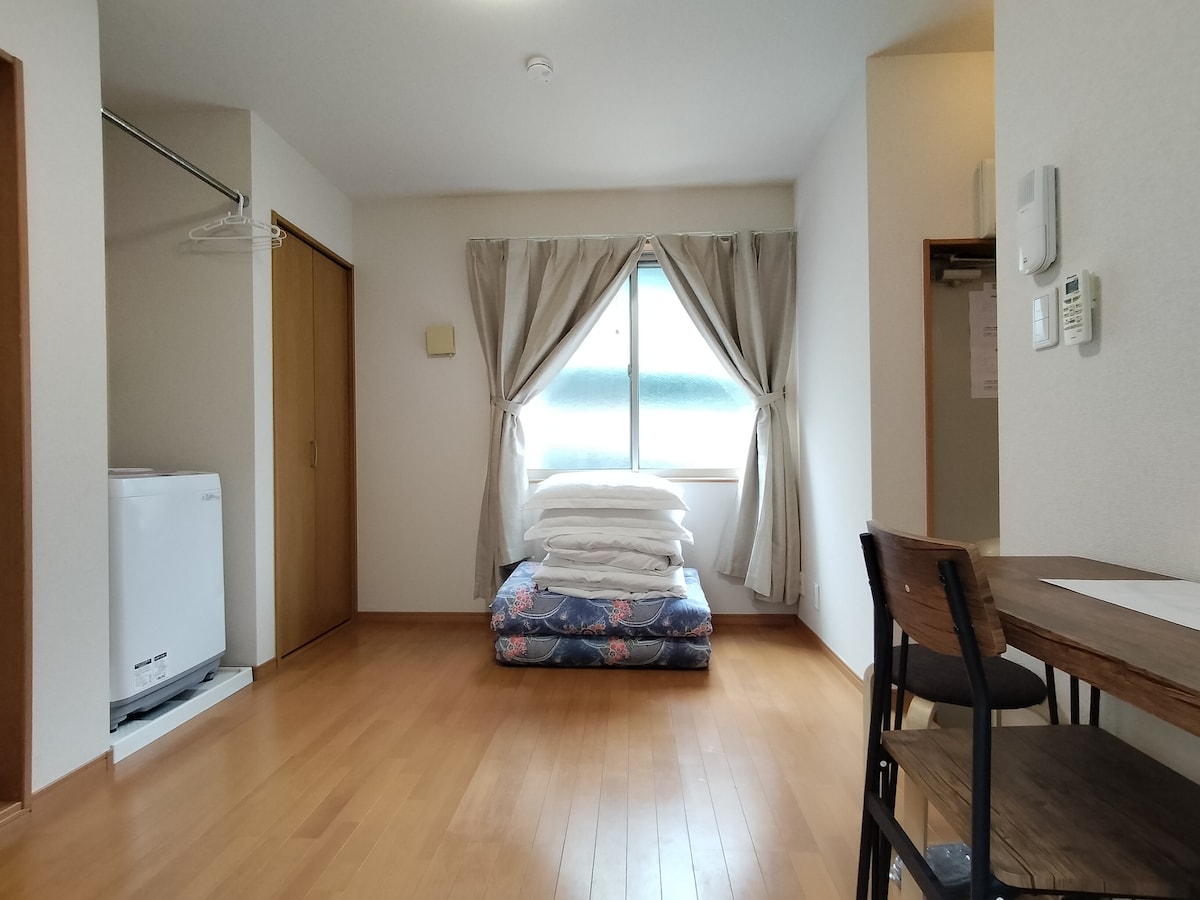 A bright room features a traditional Japanese-style futon bedding set up on a patterned mattress. A large window is dressed with light curtains, allowing natural light to fill the space. A wooden table and chair are positioned nearby, with a laundry unit visible in the corner.