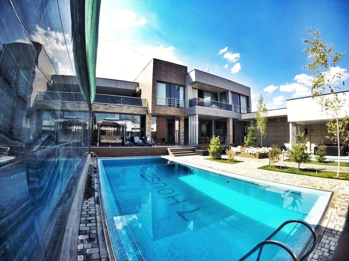A modern villa is depicted, featuring a bright blue swimming pool and sleek architecture. Reflective surfaces and large windows create a connection between indoor and outdoor spaces. Lush greenery surrounds the property, while a spacious patio with seating is visible in the background.