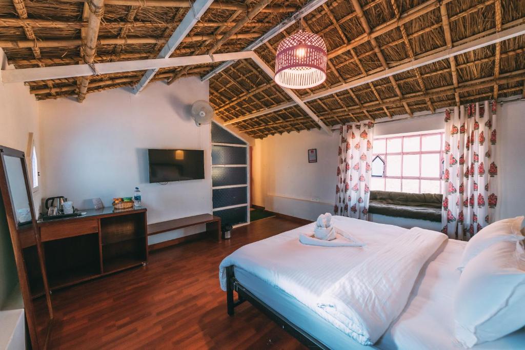 A spacious room features a thatched ceiling and rustic wooden accents. A cozy bed with white linens is centered in the space. A television is mounted on the wall opposite, with a side table and kettle visible. Large windows are dressed with floral curtains, allowing natural light.
