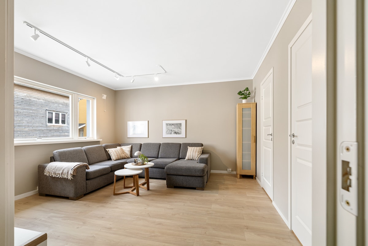 A spacious living area features a grey sectional sofa with cushions, complemented by a round coffee table. Natural light filters through large windows, illuminating the neutral-colored walls. A tall glass cabinet and decorative frames complete the inviting space.