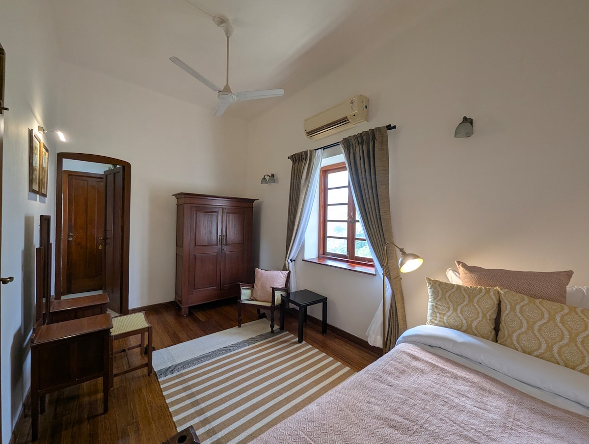 The Coral bedroom features a spacious bed adorned with neutral-toned linens. A wooden wardrobe and small table are positioned thoughtfully against the wall. Natural light enters through the window, complemented by soft curtains, creating a serene atmosphere.
