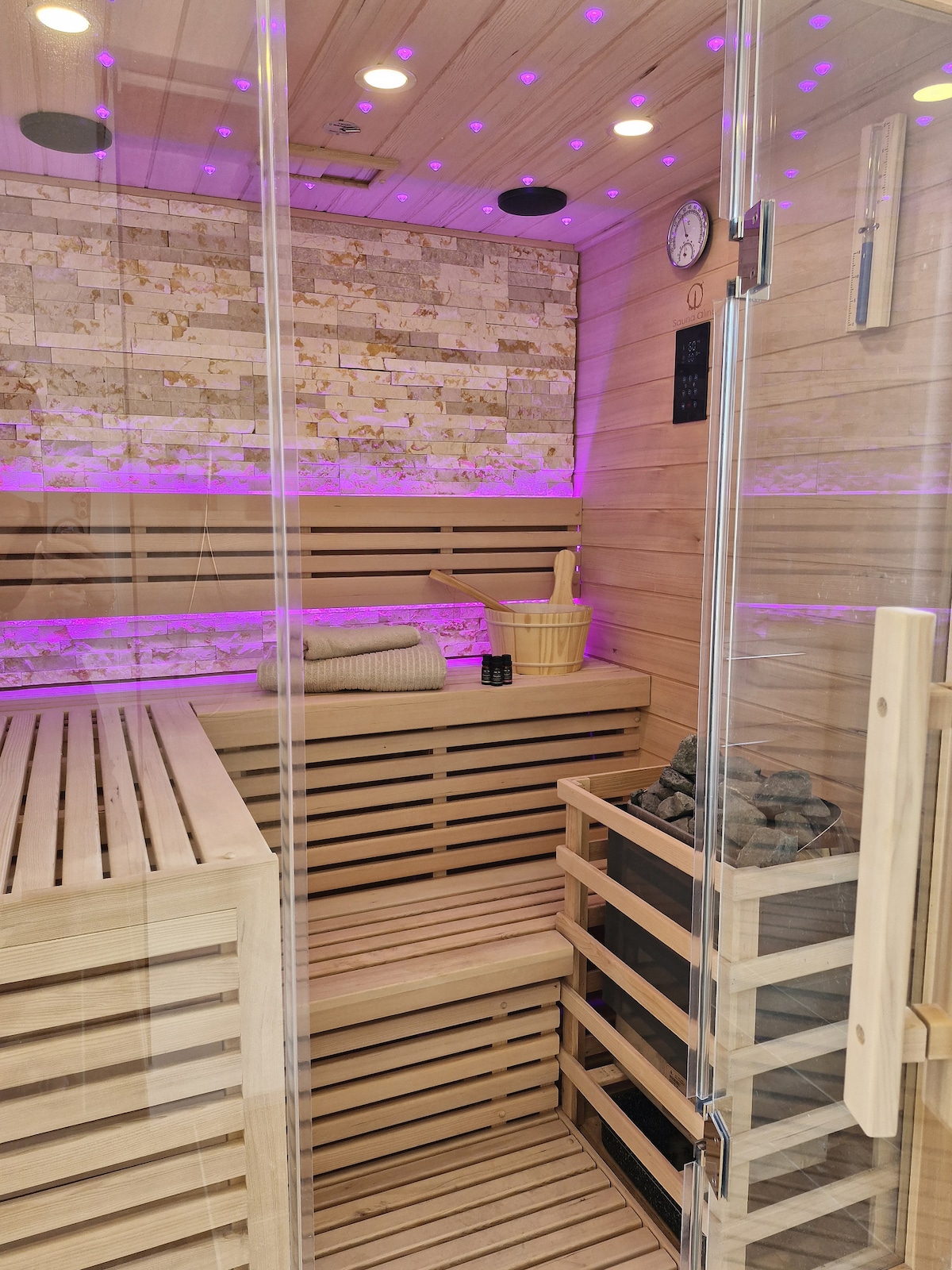 A traditional sauna is presented with warm wooden benches and stone features. Soft purple lighting enhances the ambiance. Accessories such as a towel and a wooden bucket are placed on the benches, contributing to a cozy and inviting space designed for relaxation.