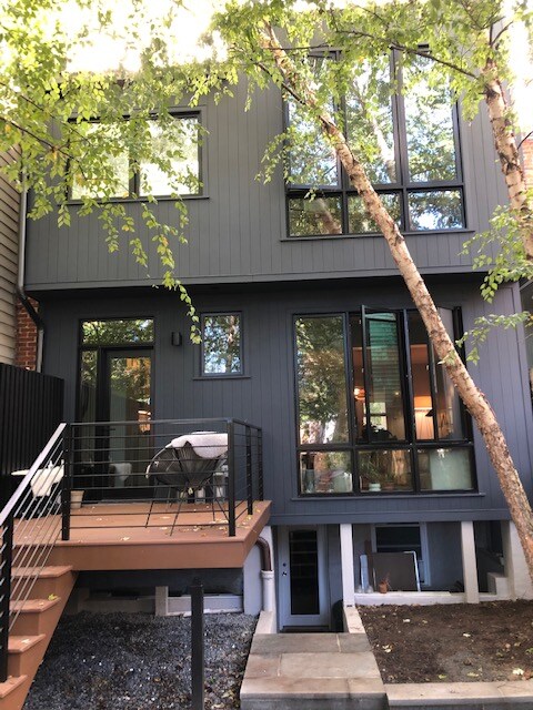 A modern exterior of a basement apartment is shown, featuring a spacious wooden deck with a seating area. Large windows provide natural light and views into the interior, while trees offer some shade. The entrance is accessible via a staircase leading down from the upper level.
