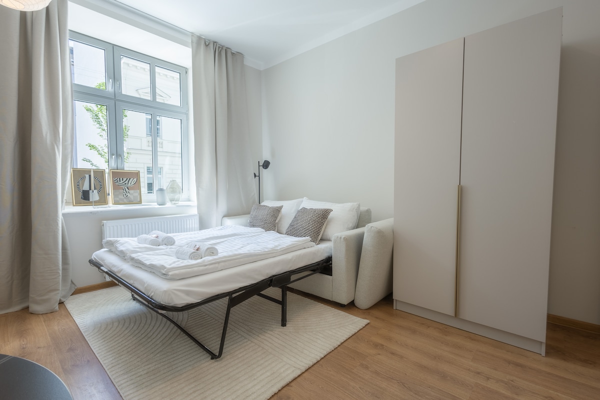 A functional living space features a folded-out sleeping area with neatly arranged linens and towels. A cozy sofa sits nearby, complemented by a large wardrobe. Natural light enters through a set of windows adorned with light curtains, brightening the neutral decor.