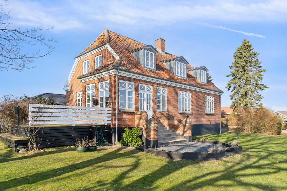 The exterior of a charming villa is displayed, featuring a brick facade with multiple windows. A large garden surrounds the property, showcasing lush grass and a variety of greenery. A spacious deck with seating options sits to the side, enhancing the outdoor area.