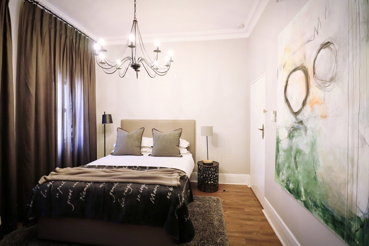 A cozy bedroom is presented with a spacious bed adorned with decorative pillows and a textured throw blanket. A modern chandelier hangs above, and two bedside lamps provide soft lighting. A large wall art piece in calming colors complements the neutral decor.
