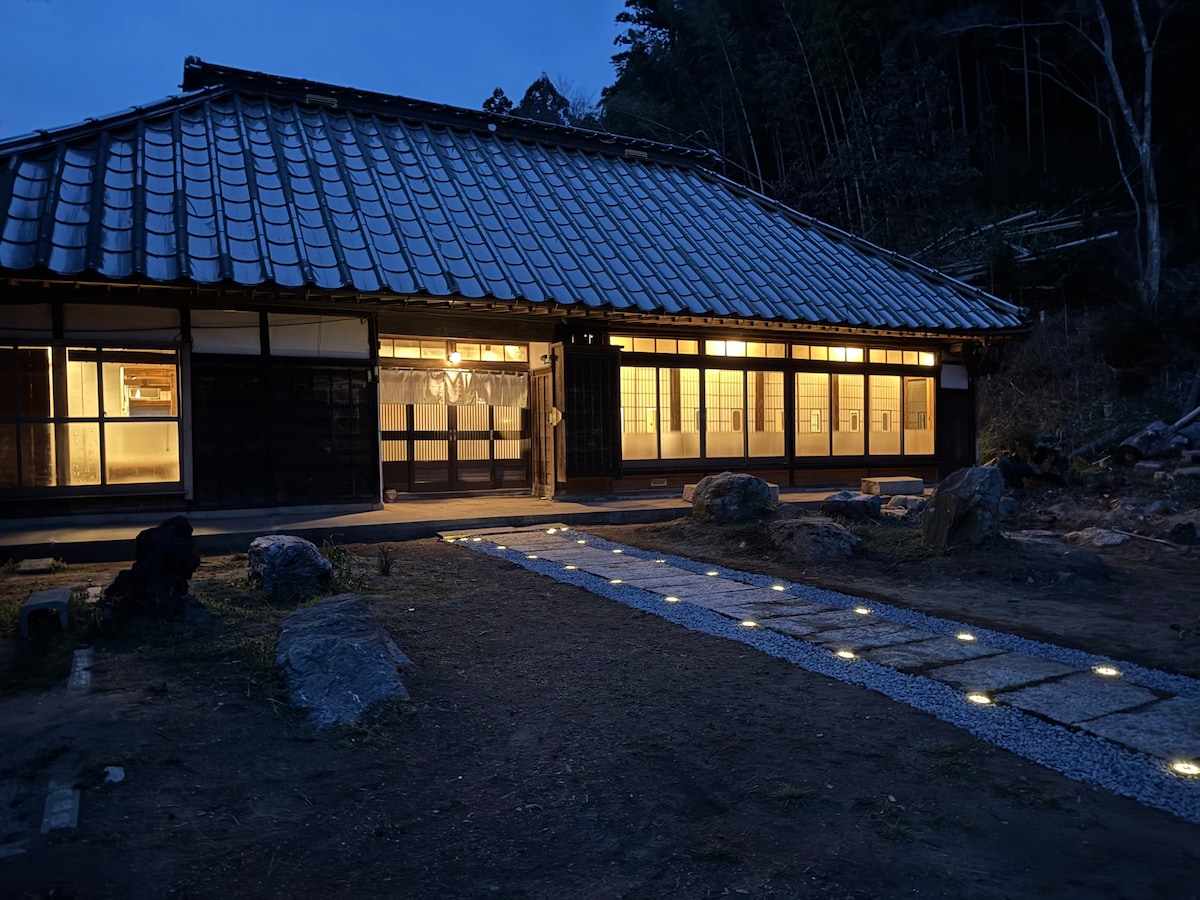 The exterior of a traditional Japanese house is illuminated at dusk, with warm light spilling from large windows. A stone pathway lined with soft ground lights leads to the entrance, surrounded by natural elements and rocky features, enhancing the serene evening ambiance.