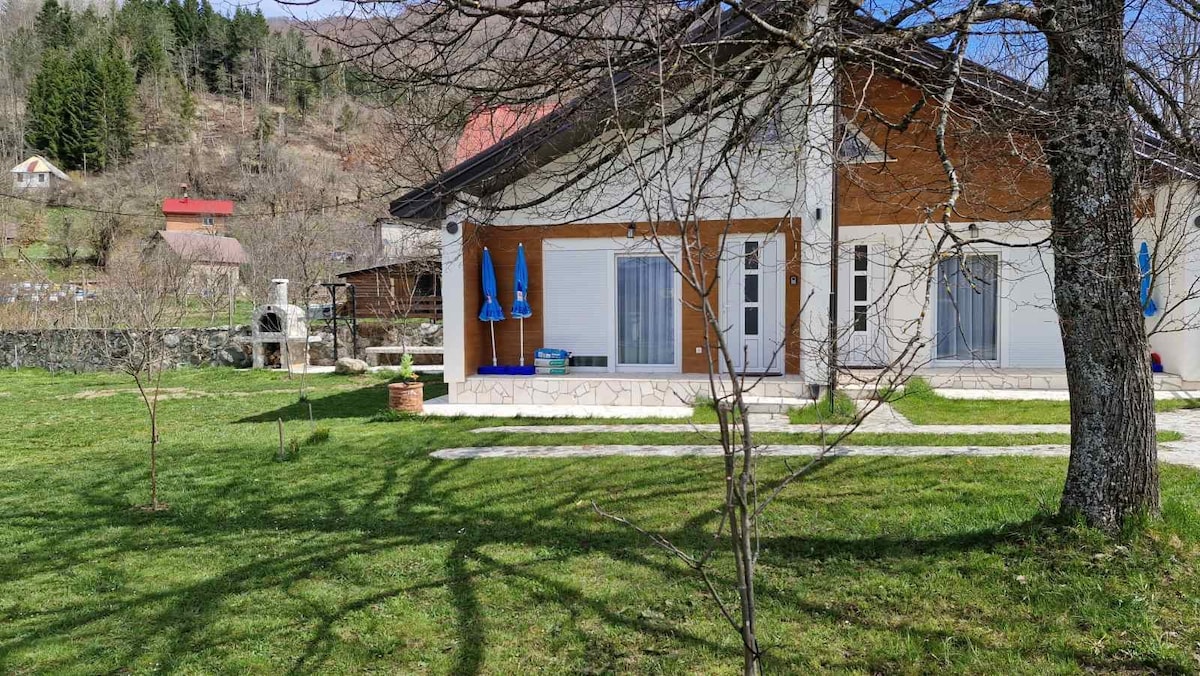 A modern two-story house is set against a backdrop of gently sloping hills. Light-colored walls are complemented by wooden accents. A spacious patio area features chairs, inviting relaxation, while a tree provides shade. Manicured grass surrounds the home, enhancing the serene outdoor space.