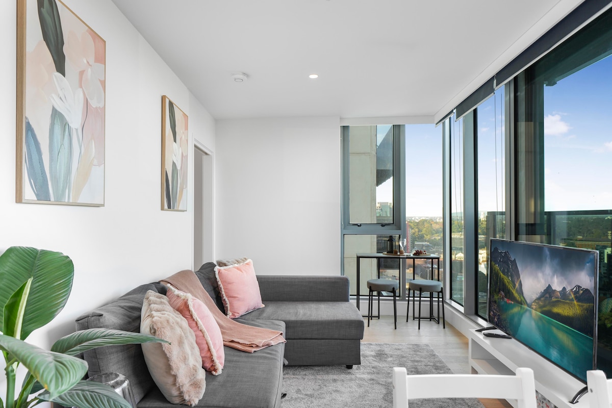 A modern living area features a comfortable gray sofa adorned with soft pillows. A wall-mounted television stands opposite, while floor-to-ceiling windows provide ample natural light and city views. A small dining table for two is visible beside the window, alongside indoor greenery.