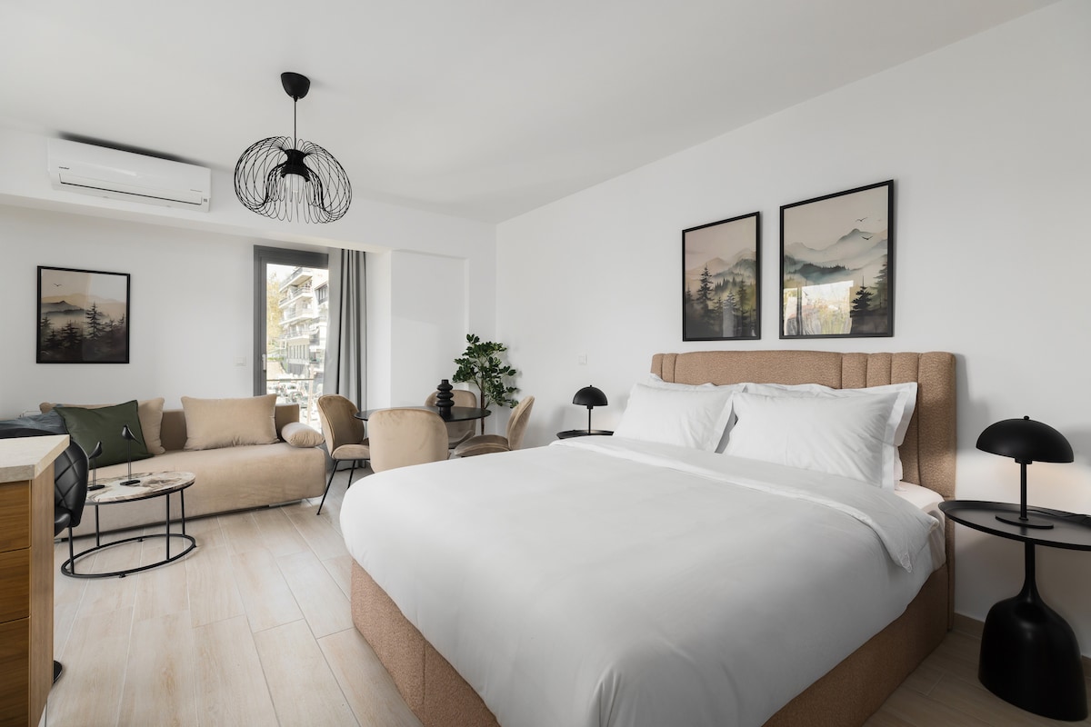 A spacious bedroom features a large, upholstered bed with crisp white linens. A cozy seating area includes a sofa, a contemporary light fixture, and artwork on the walls. Natural light enters through glass doors that lead to a balcony, enhancing the room's airy feel.