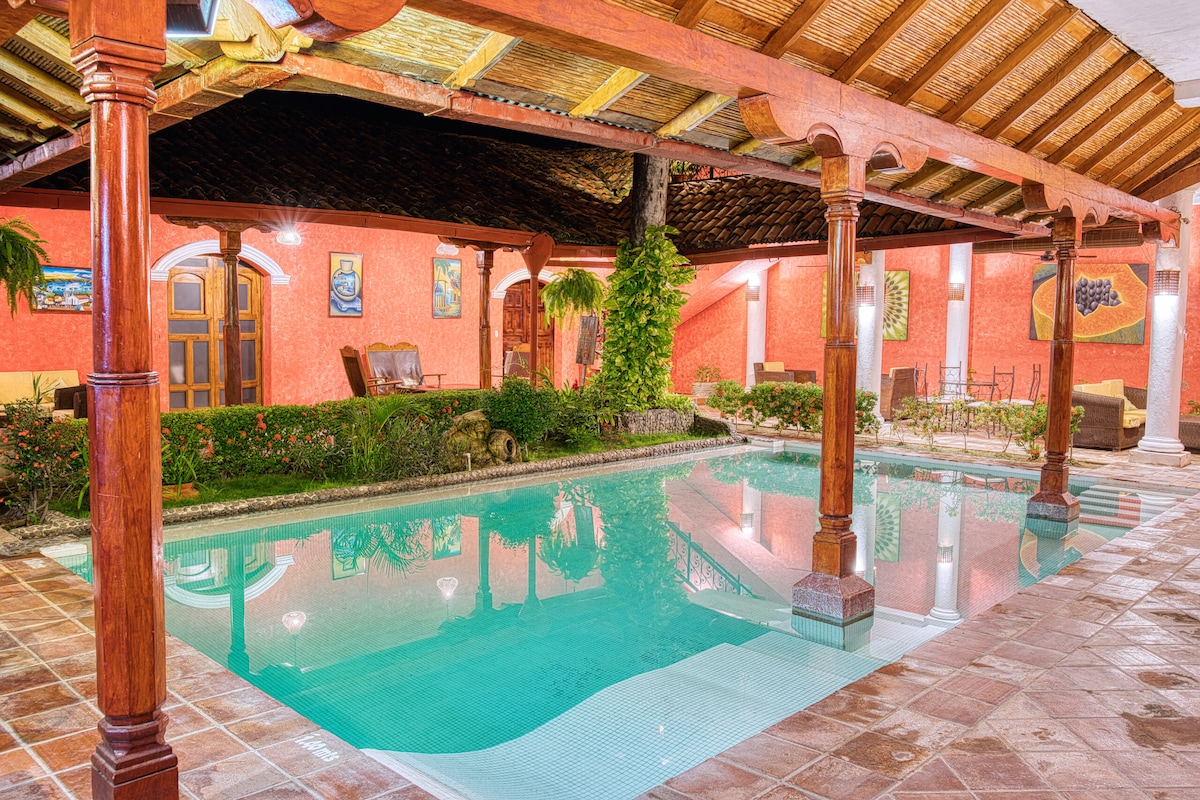 A well-maintained outdoor pool is surrounded by a spacious terrace, with wooden beams overhead providing shelter. Lush greenery and vibrant artwork are positioned around the area, creating an inviting atmosphere. Comfortable seating options can be seen, enhancing the space for relaxation.