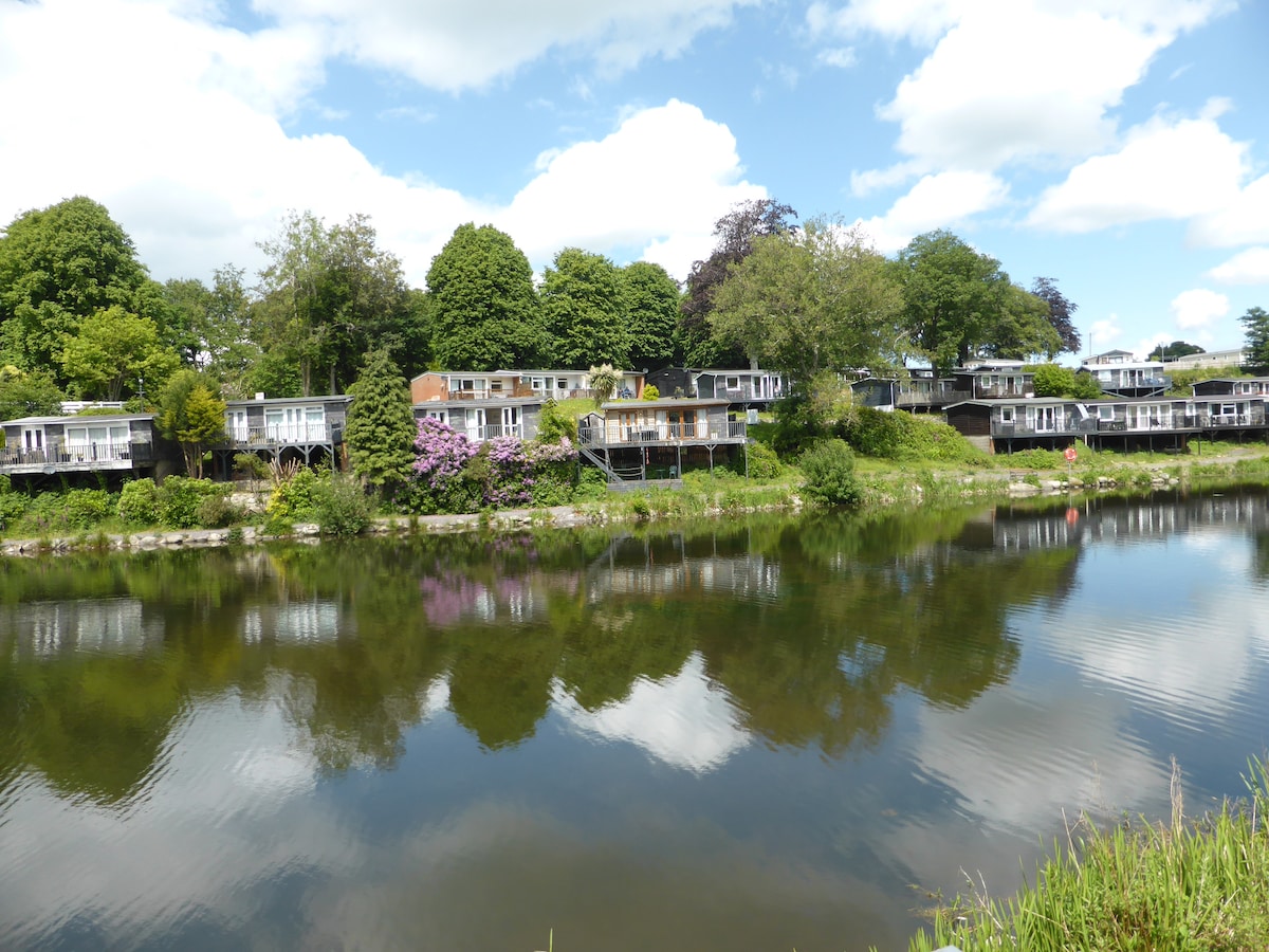 A row of cabins is situated along the edge of a serene lake, surrounded by lush greenery. Reflections of the cabins and trees can be observed in the calm water. The sky is bright with scattered clouds, enhancing the peaceful atmosphere.