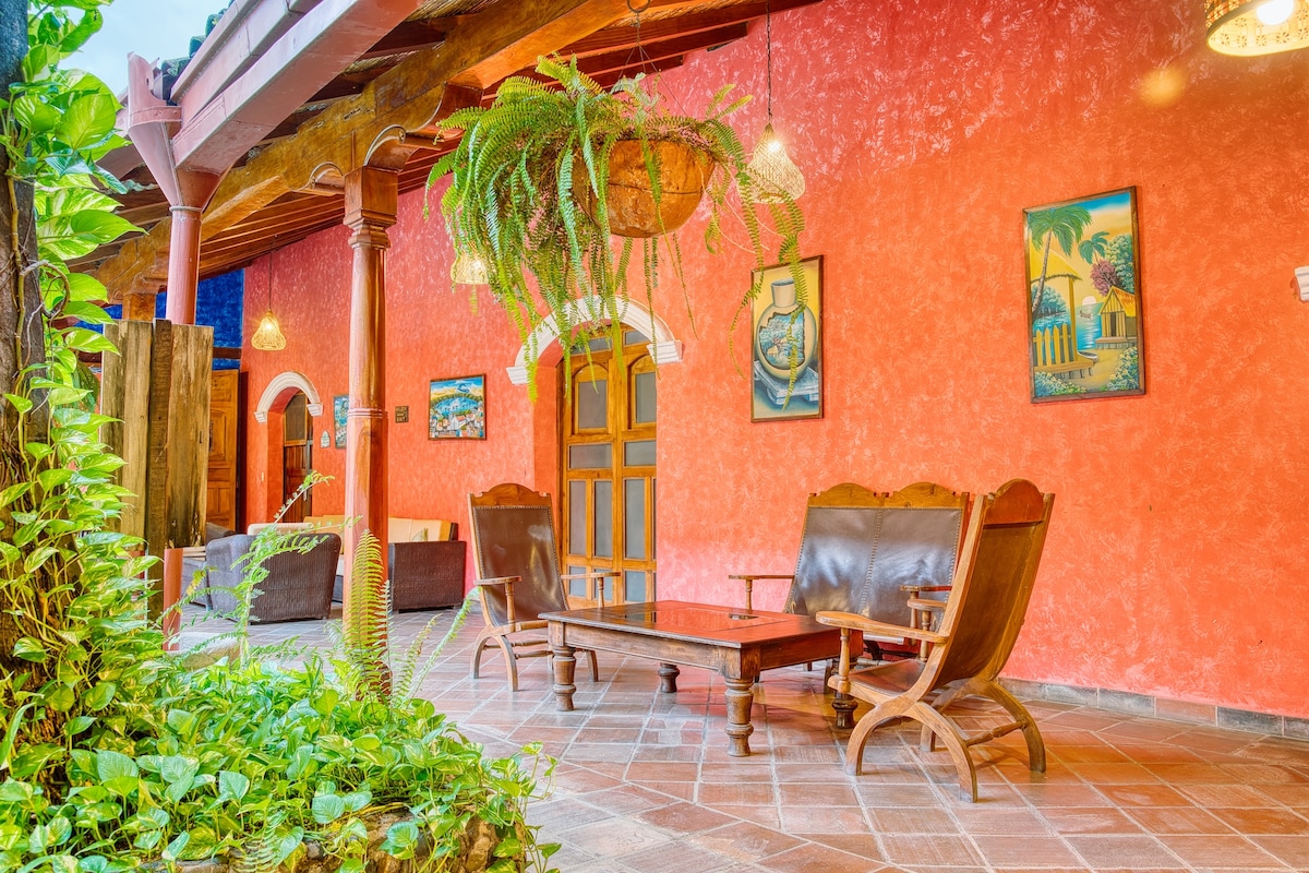 A vibrant common area displays rustic wooden furniture against a bright red wall. Potted plants hang from the ceiling, adding greenery. Decorative art adorns the walls, while soft lighting creates a relaxed atmosphere. The tiled floor enhances the inviting space for gathering.