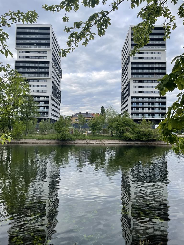 Two modern high-rise buildings are reflected in the calm water of a river, surrounded by lush greenery. The architecture features multiple balconies and large windows, enhancing the contemporary appeal of the structures. A partly cloudy sky is visible above, providing a serene backdrop.