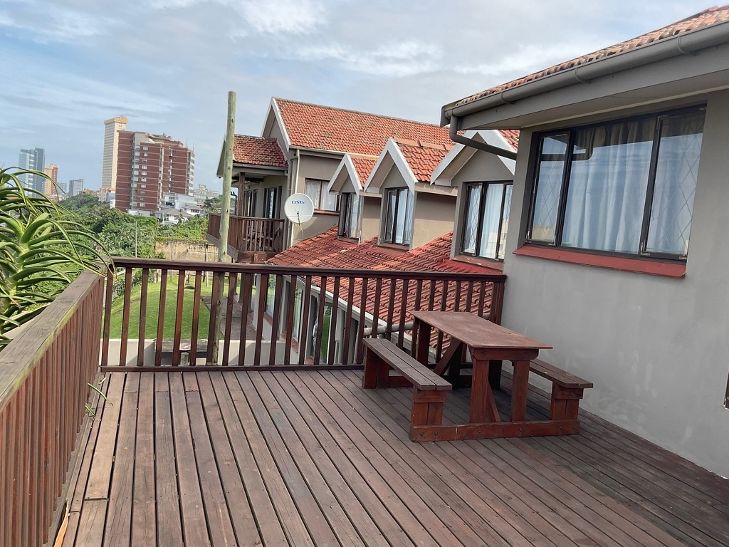 A wooden deck is featured, with a picnic table and benches inviting casual gatherings. The background displays multiple rooftop structures, while a clear sky sets a calm atmosphere. Lush greenery is visible adjacent to the deck, enhancing the setting's natural appeal.