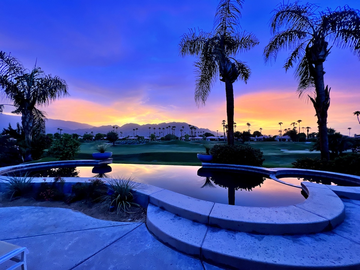 A tranquil view of the golf course is captured at sunset, with vibrant hues of orange and purple reflecting off a black-bottom infinity edge pool. Lush palm trees and greenery frame the scene, enhancing the serene atmosphere.