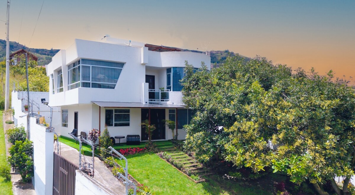 A modern house is surrounded by lush greenery and flowering plants. Large windows dominate the facade, offering views of the serene landscape. The entrance features a spacious porch with seating, while a well-maintained yard slopes gently toward the driveway.