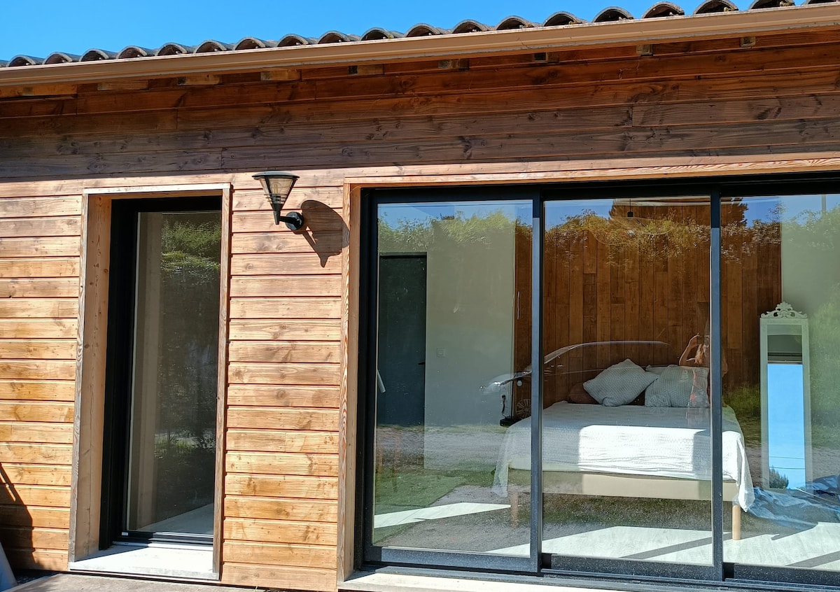 A wooden exterior reflects a natural aesthetic, featuring large glass doors that provide a view of a bright sleeping area with a spacious bed. The entrance is framed by two separate doors, emphasizing the independent access to the studio.