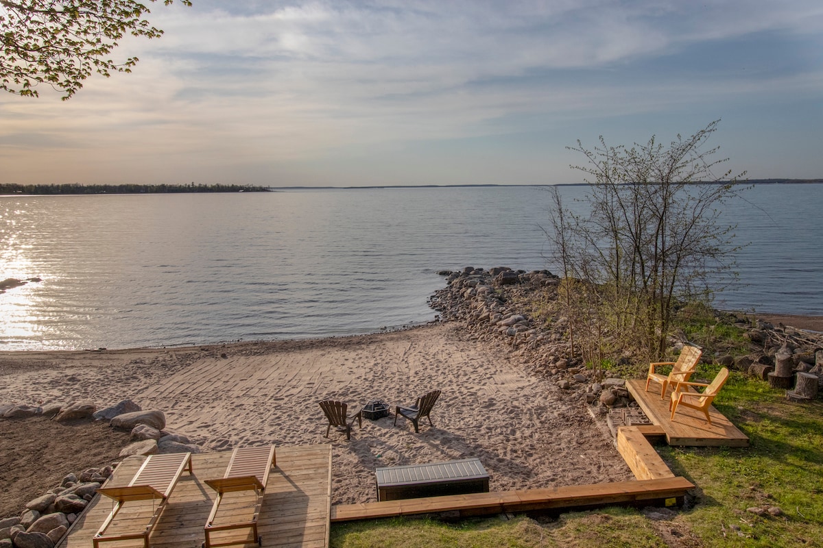 A sandy beach area overlooks tranquil water, with smooth stones lining the shore. Two wooden lounge chairs and a firepit with seating are positioned on the sand. A wooden deck extends towards the water, providing an inviting spot to enjoy picturesque views.