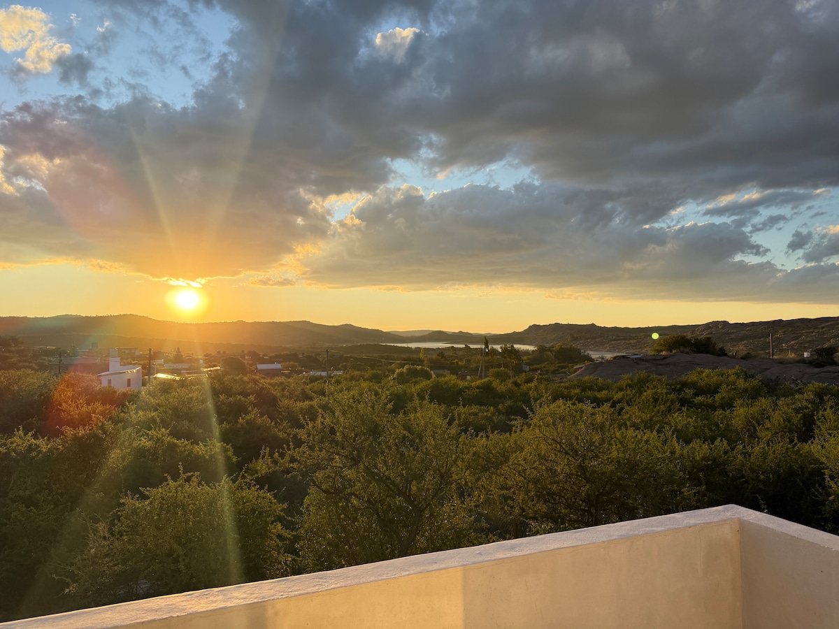 A vibrant sunset casts warm hues over the landscape, revealing a scenic view of the surrounding greenery and the lake in the distance. The sky is filled with soft oranges and yellows, creating a serene atmosphere at dusk.
