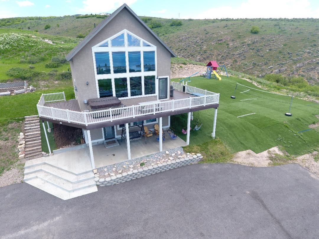 A spacious lakeside house features a large front deck with seating arrangements. Expansive windows provide natural light and scenic views. The yard includes a grassy area, a climbing playset, and open space for outdoor games, surrounded by gentle hills and greenery.