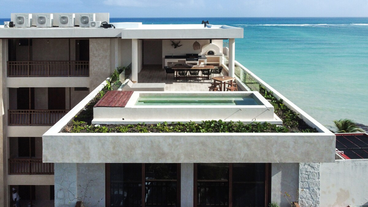 A rooftop area presents an infinity plunge pool surrounded by lush greenery. Dining and lounge spaces are situated adjacent to the pool, with a panoramic view of the ocean visible in the background. The design is modern, harmonizing with the natural surroundings.