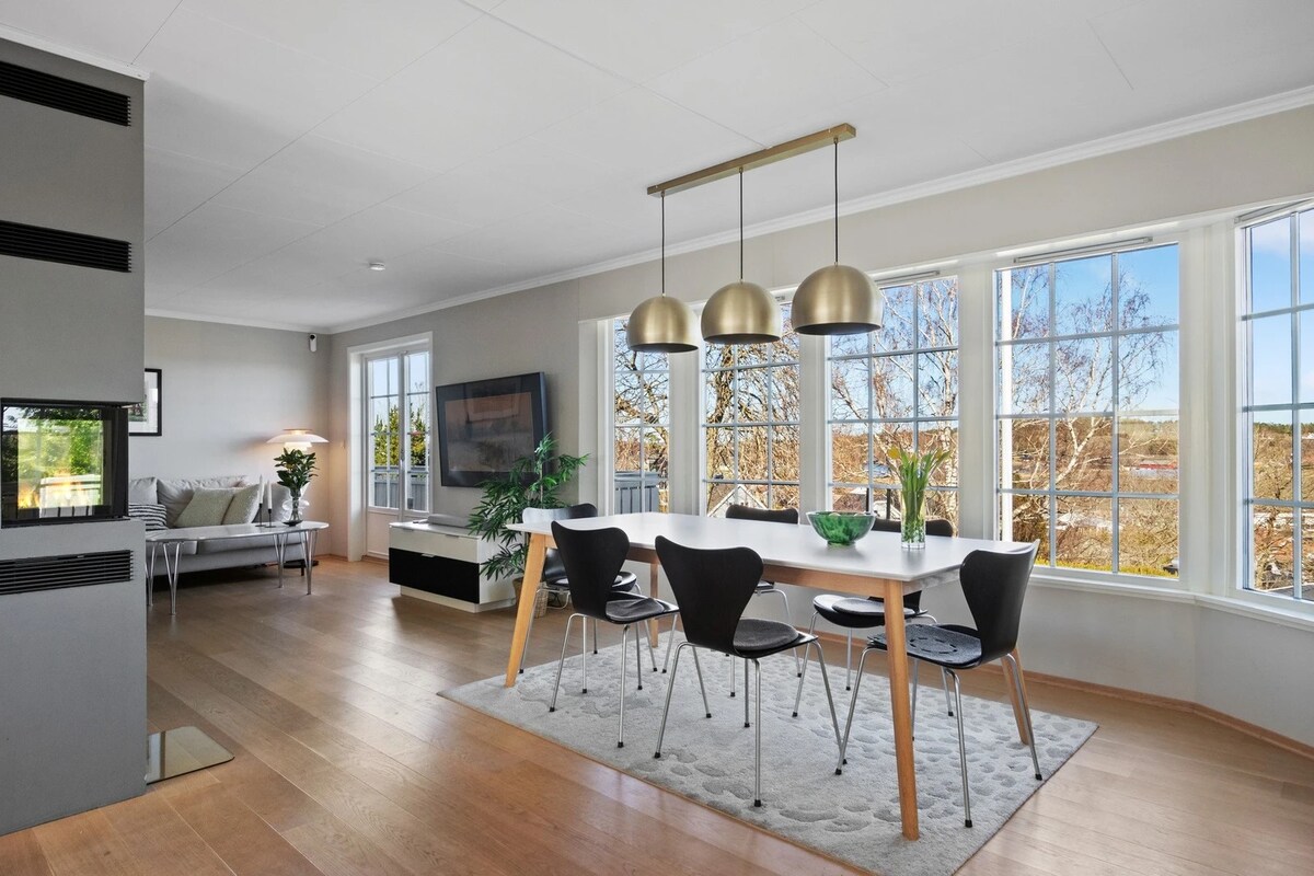 A bright and airy open-plan living space is visible, featuring large windows that invite natural light. A modern dining table with four chairs is centered within the room, complemented by stylish pendant lights above. A comfortable seating area is positioned to the side with greenery enhancing the ambiance.