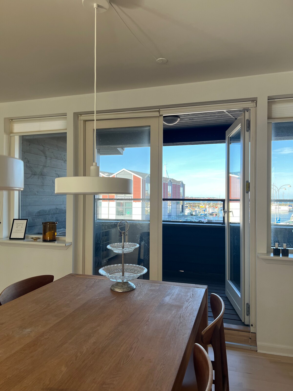 A dining area features a wooden table accompanied by elegant chairs. Bright natural light enters through large glass doors that open to a balcony overlooking the harbor and city. Simple decor elements are arranged on the table for a modern touch.